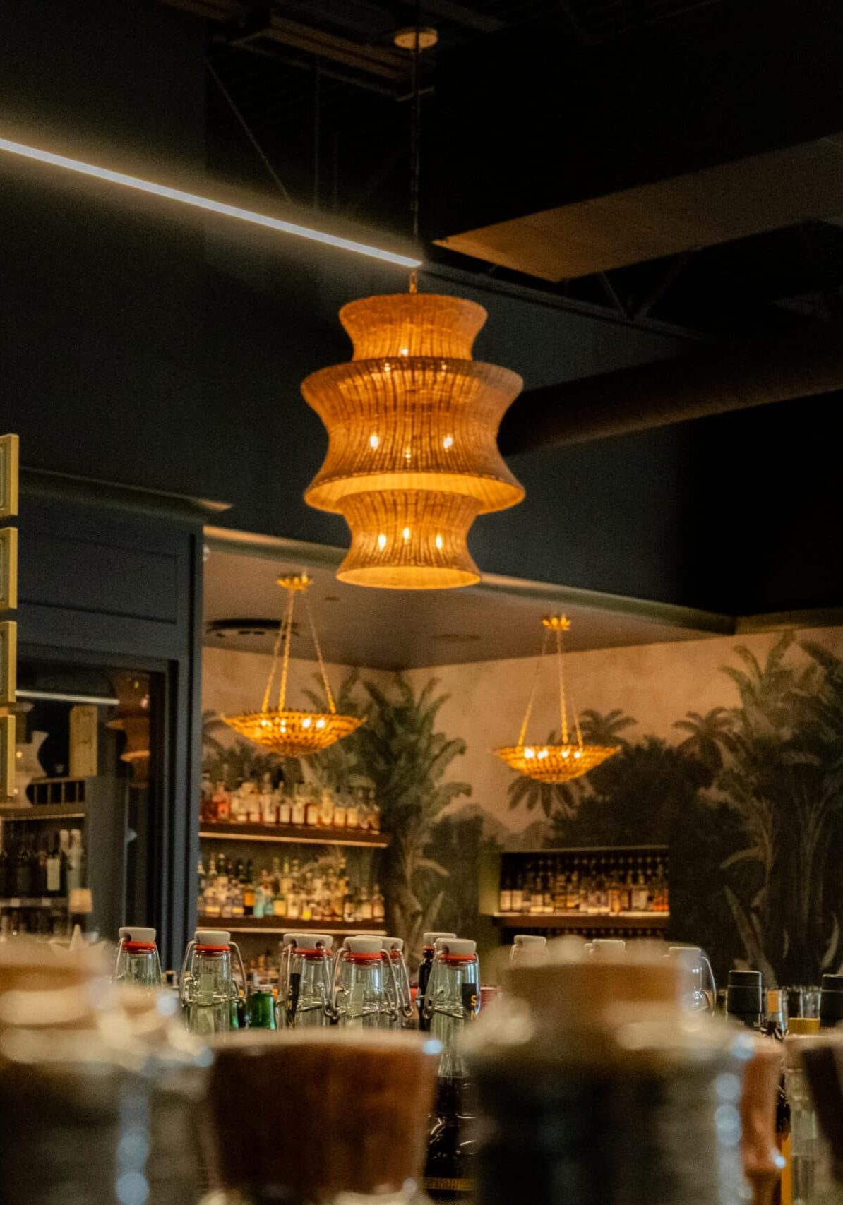 Bar interior with assorted bottles on the counter, hanging chandeliers, decorative signage, and a tropical-themed mural on the back wall.