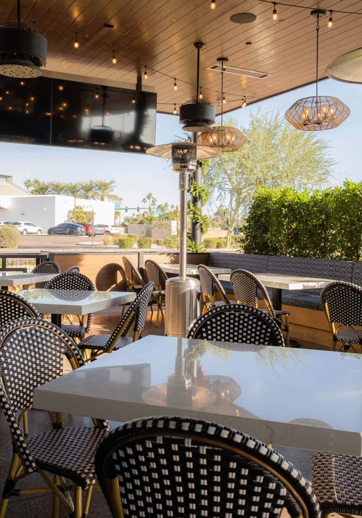 Outdoor restaurant patio with empty tables and chairs, patterned cushions, potted plants, hanging lights, and a large TV, with a street and buildings visible in the background.
