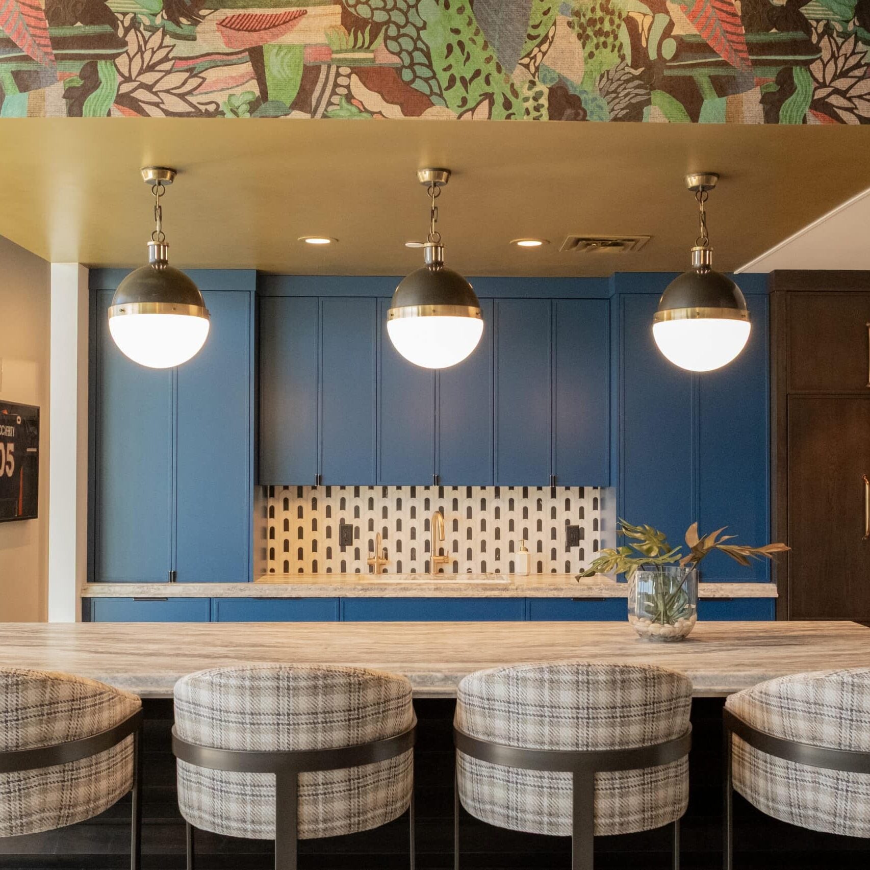 Modern kitchen with a patterned ceiling, blue cabinets, hanging globe lights, a large island with plaid barstools, and a vase with greenery on the countertop.