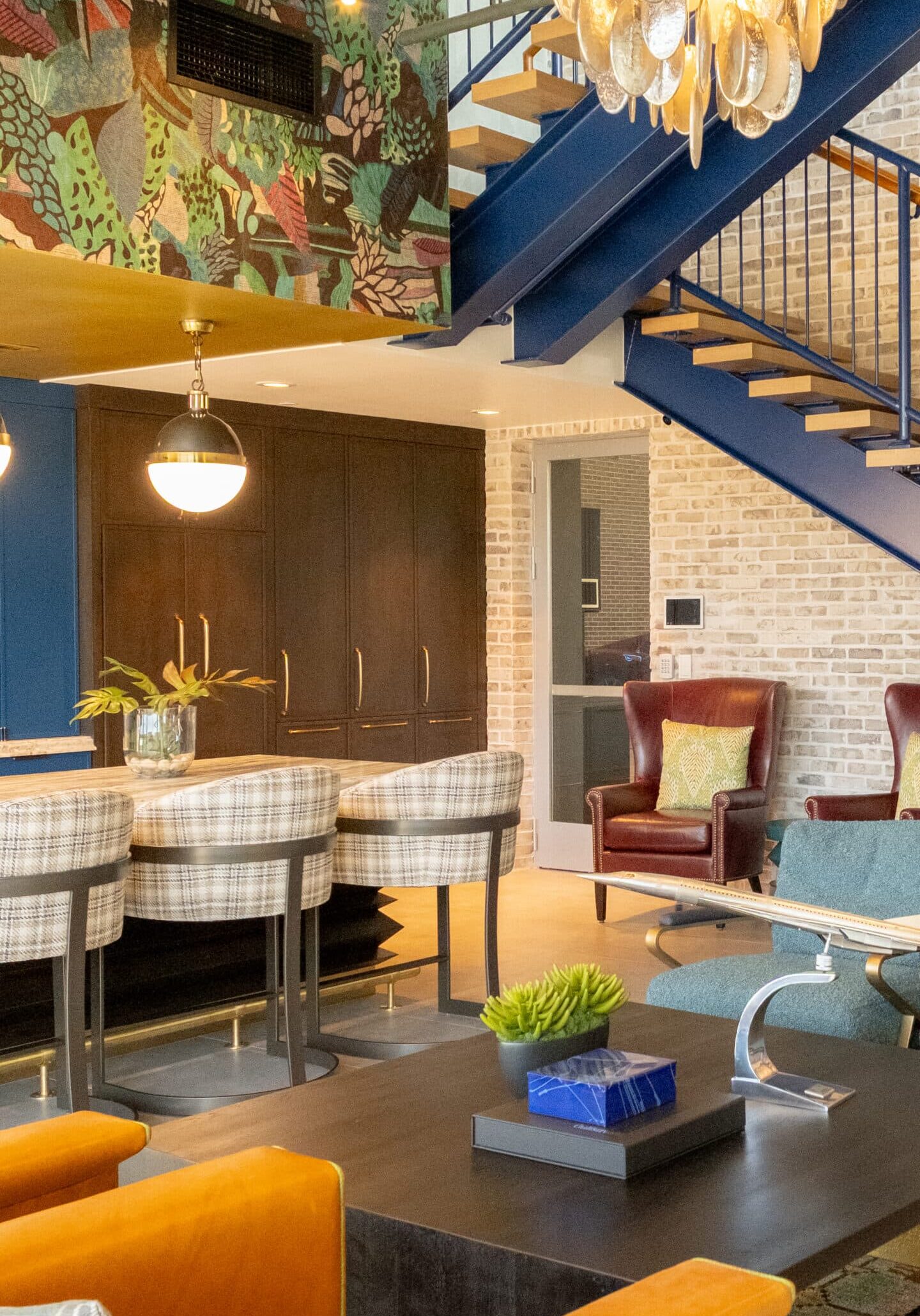 Colorful modern living space with orange sofa, plaid bar stools, leather armchairs, large chandelier, patterned wall art, and open blue staircase against exposed brick.