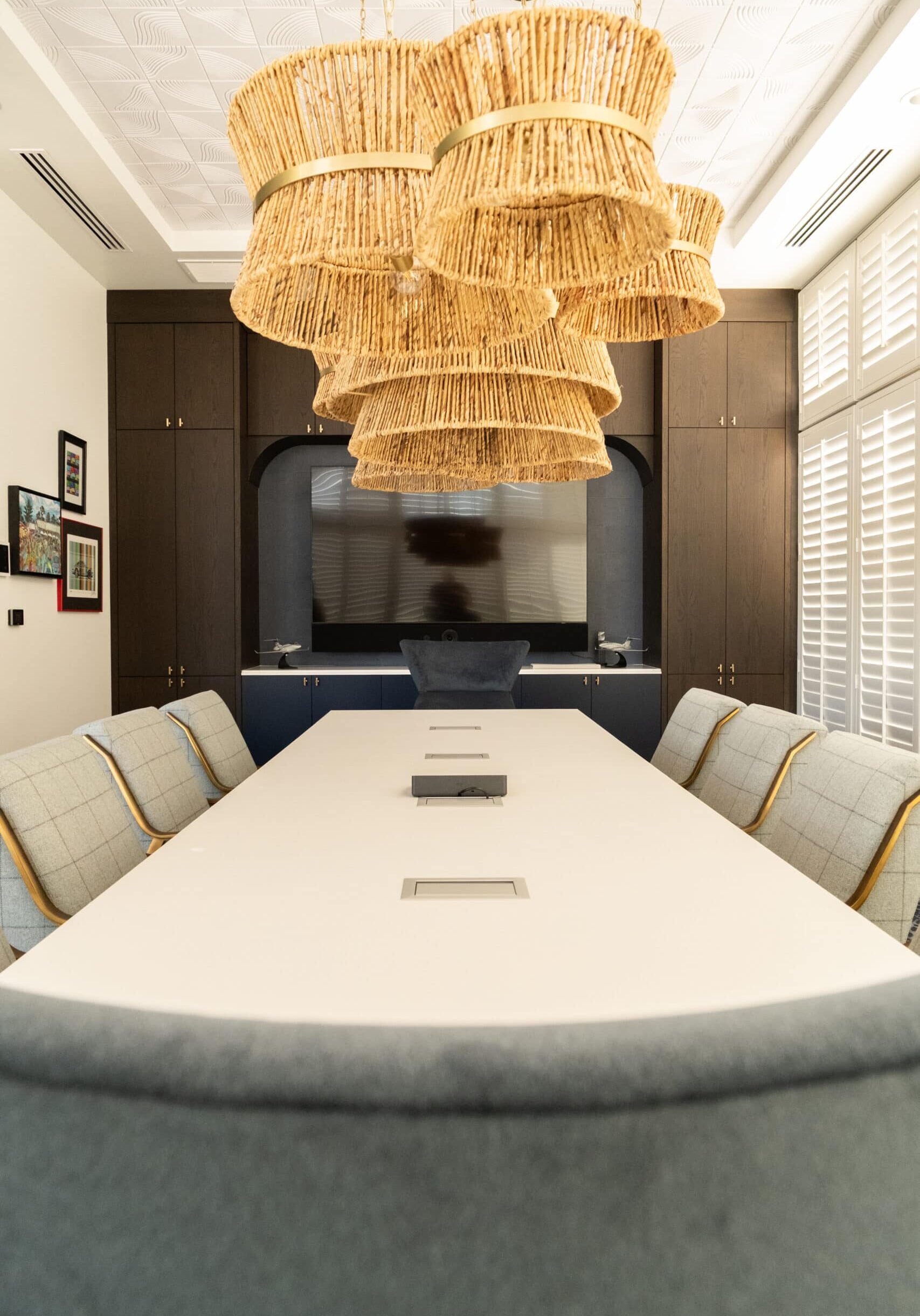 A modern conference room with a long white table, cushioned chairs, woven pendant lights, and a large television on a dark built-in cabinet.