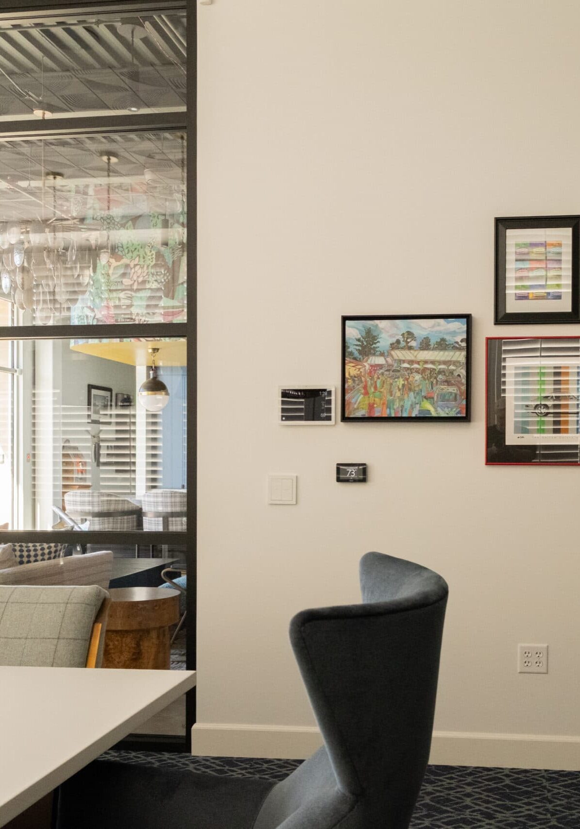 A modern office space with a white table, gray chairs, wall art, a large woven light fixture, and a glass wall revealing a lounge area and parked cars outside.