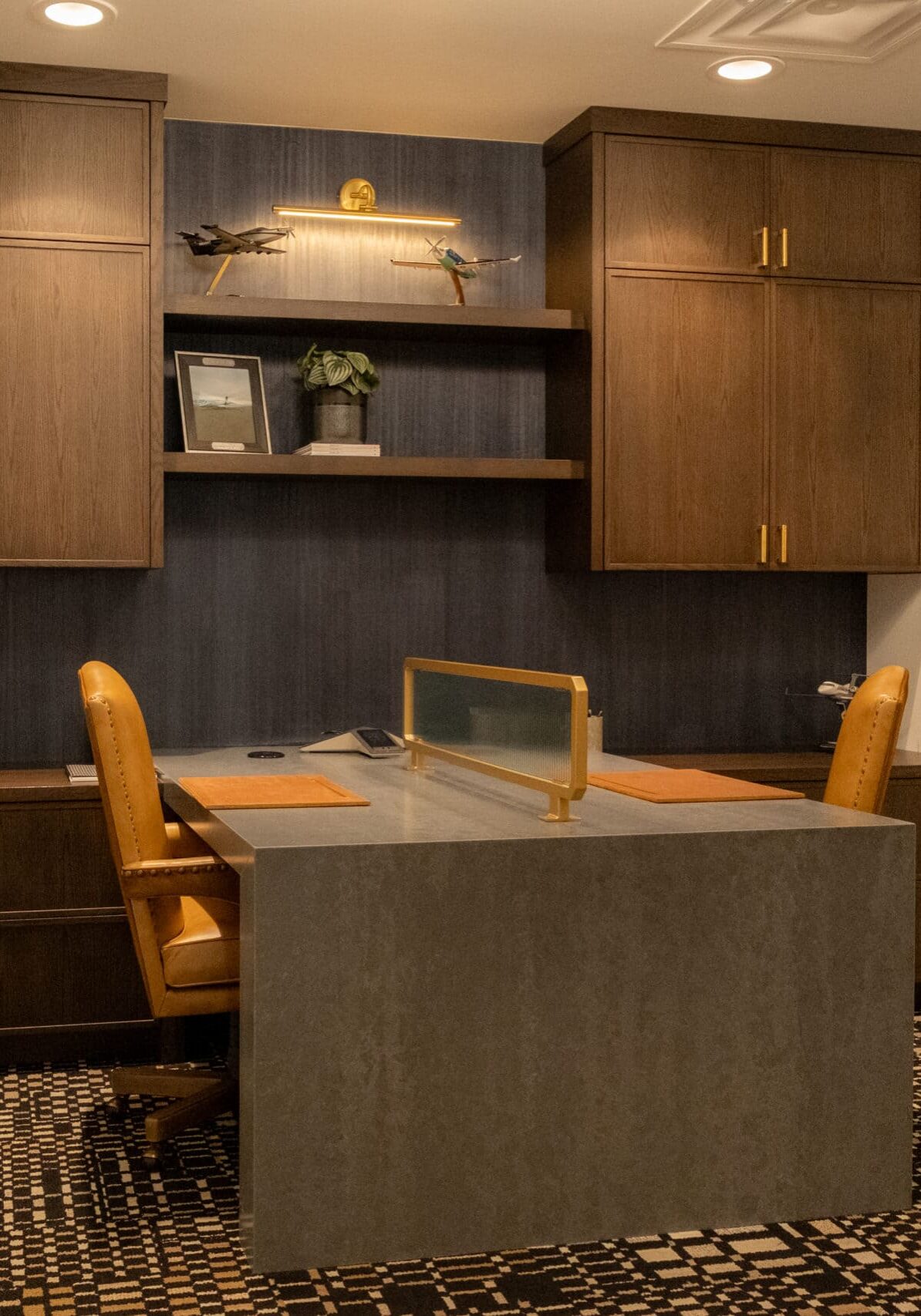 Modern home office with a gray desk, two tan chairs, wooden cabinets, framed photos on the wall, and a decorative airplane model on a shelf.