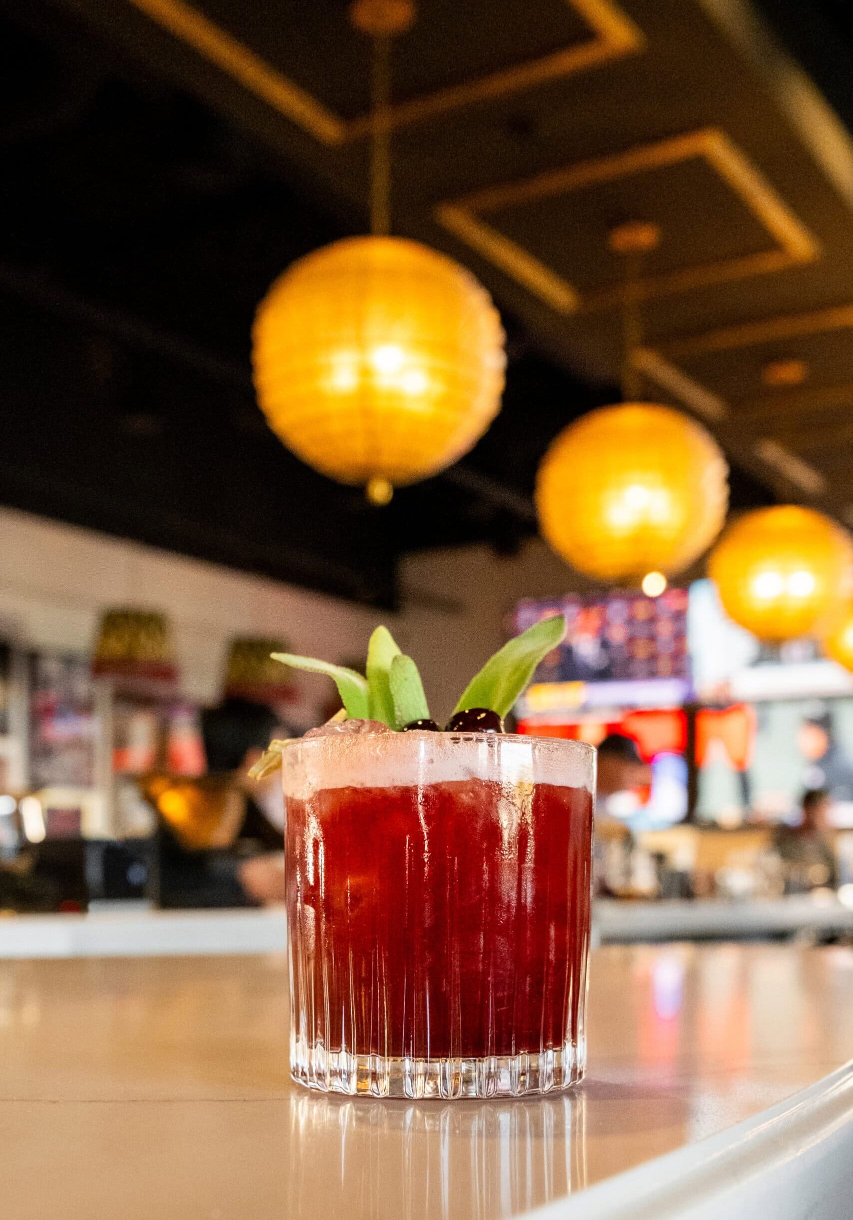 A red cocktail garnished with green leaves and a cherry sits on a bar counter, with warm, round pendant lights hanging in the background.