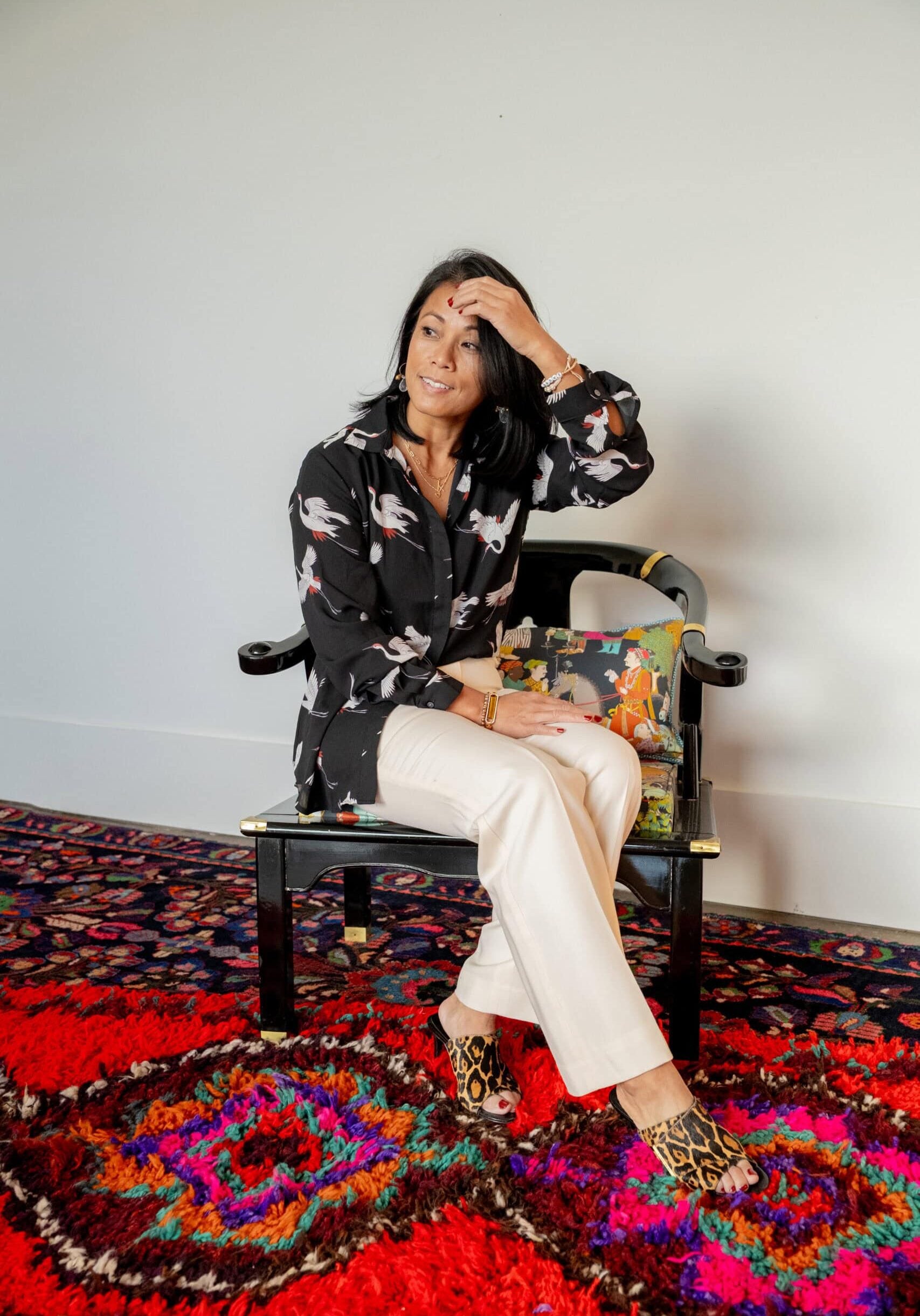 A woman with dark hair sits on a black chair with a colorful cushion, wearing a black patterned blouse, light pants, and leopard print shoes, on a red patterned rug.