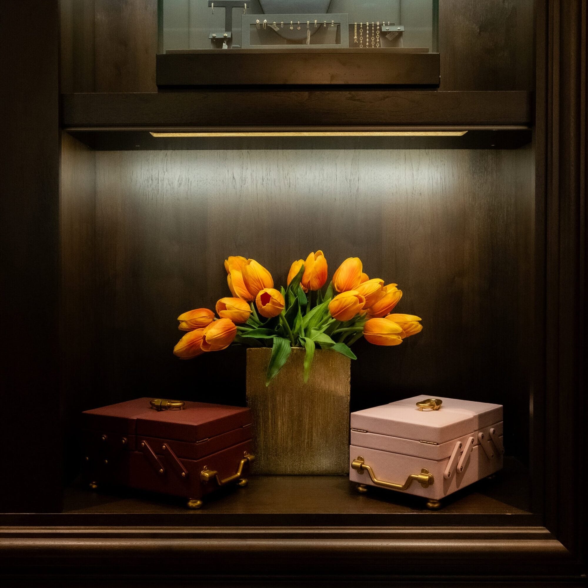A vase of orange tulips is placed between a closed maroon box and a closed pink box, with jewelry displayed on a shelf above.