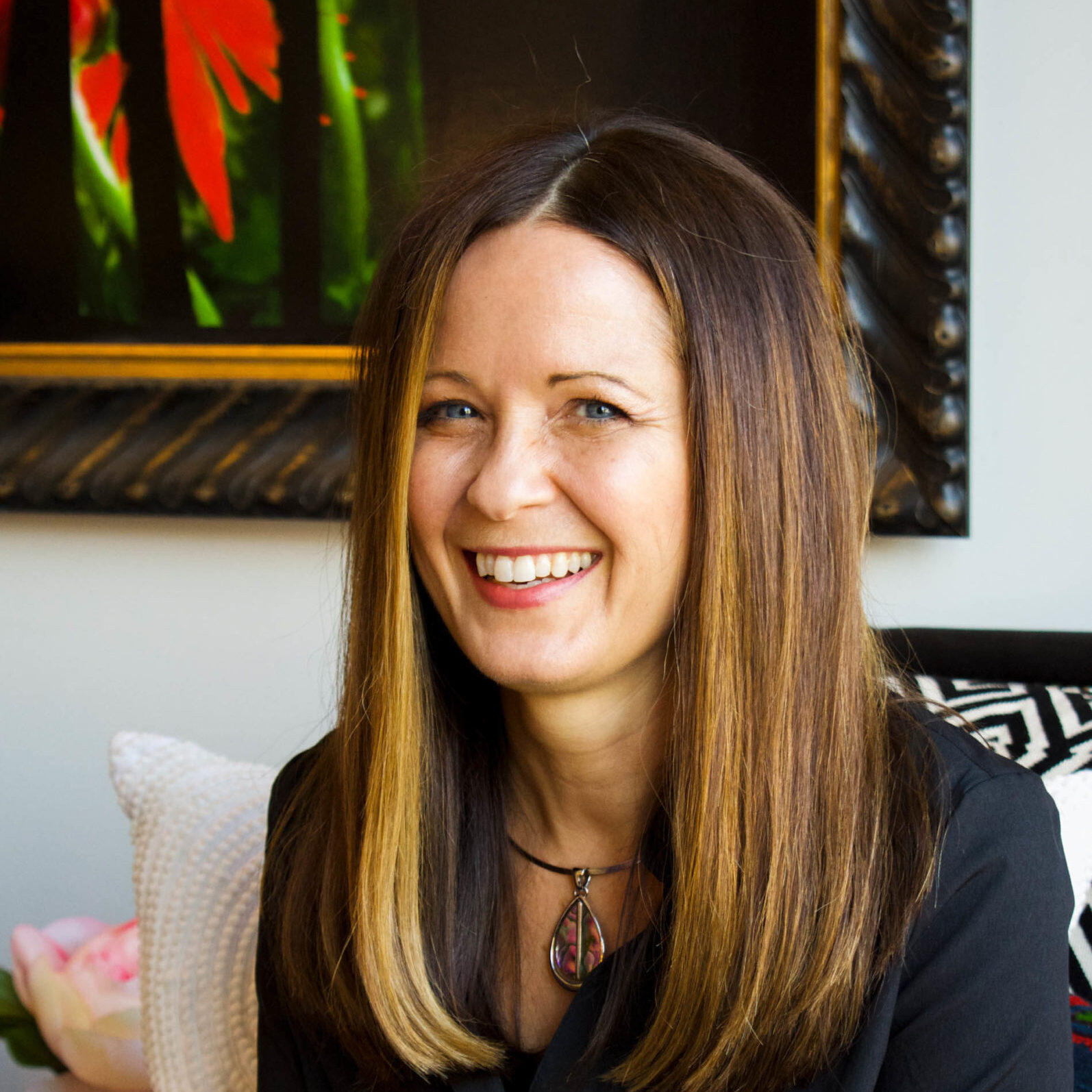 A woman with straight, long brown hair and a necklace smiles while sitting indoors in front of a framed, colorful painting—a vibrant interior reminiscent of Crimson Design Group’s signature style.