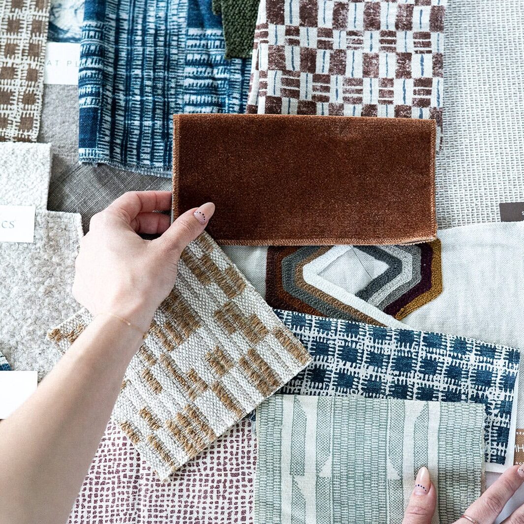 A person arranges various fabric swatches with different patterns and textures, some labeled with Crimson Design Group brand tags, on a flat surface.