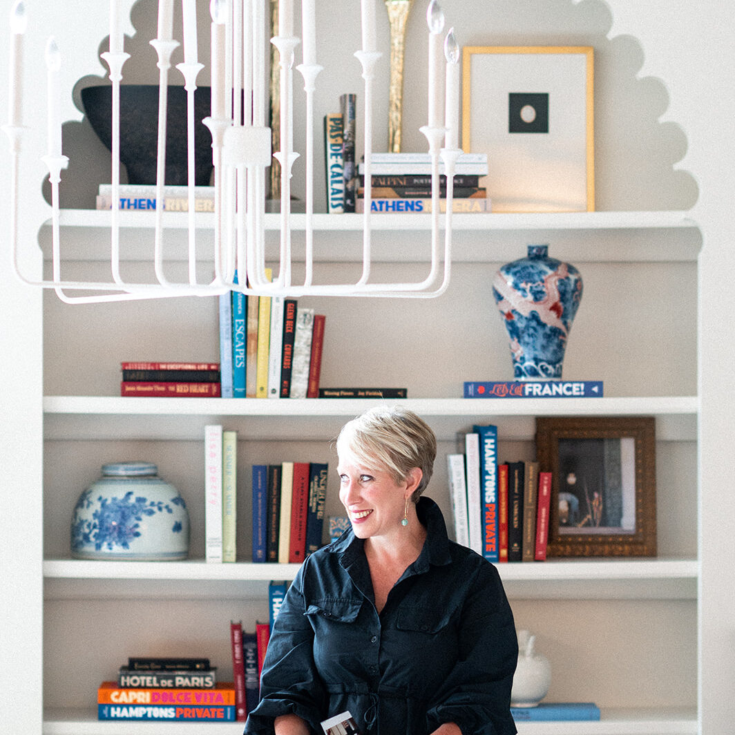 A woman with short blonde hair stands at a table, flipping through a book. Behind her, bookshelves and decorative vases showcase the signature elegance of Crimson Design Group beneath a striking white chandelier.