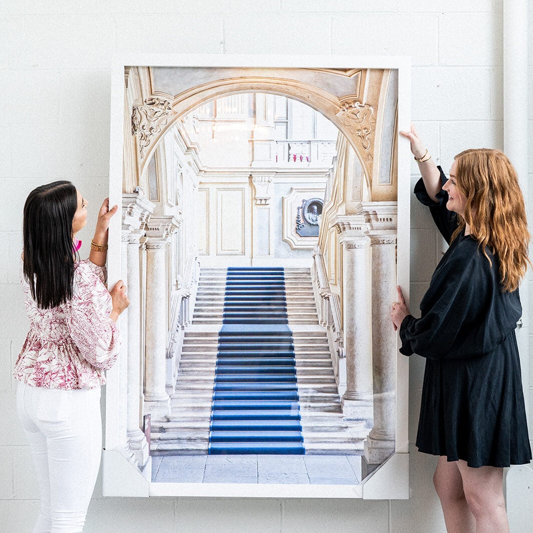 Two women stand on either side of a large framed photograph of a grand staircase with a blue carpet, mounted on a white wall—an elegant display that reflects the signature style of Crimson Design Group.
