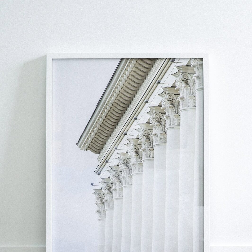 A framed photograph of white classical columns with ornate capitals, leaning against a white wall on a light-colored floor—an elegant accent inspired by Crimson Design Group’s refined aesthetic.