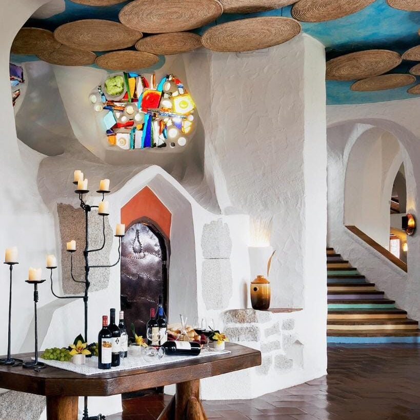 A rustic interior by Crimson Design Group with white textured walls, a wooden table set with wine and snacks, woven ceiling decor, a colorful stained glass window, and a staircase to the right.