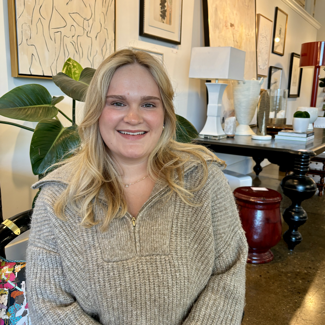 A woman with blonde hair and a beige sweater sits and smiles in a modern, art-filled room curated by Crimson Design Group, featuring plants and decorative objects in the background.