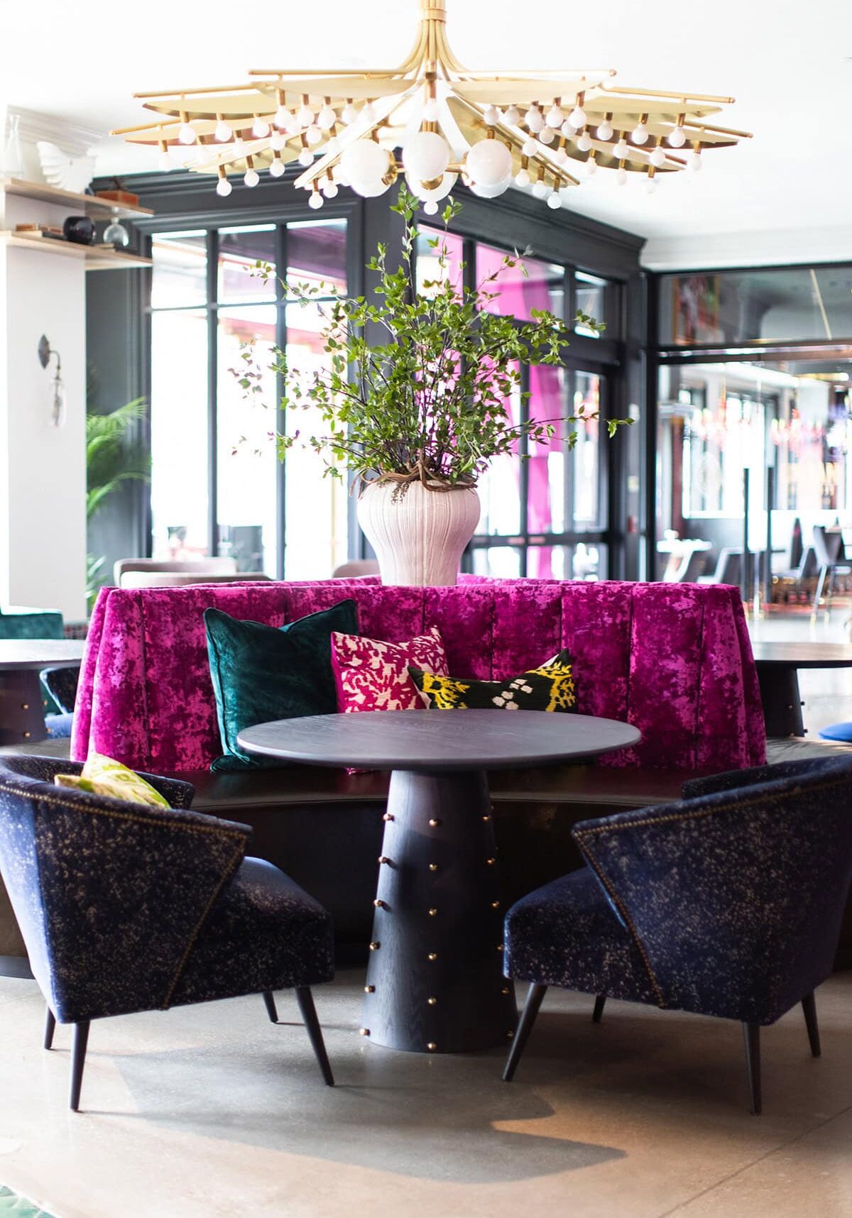 A round black table with dark blue chairs and a magenta velvet banquette, featuring assorted cushions and a large vase with green branches, under a modern chandelier.