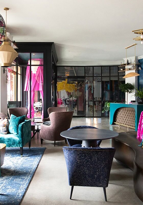 A modern lounge with colorful velvet seating, decorative pillows, large globe lamps, plants, and a mural on the wall.