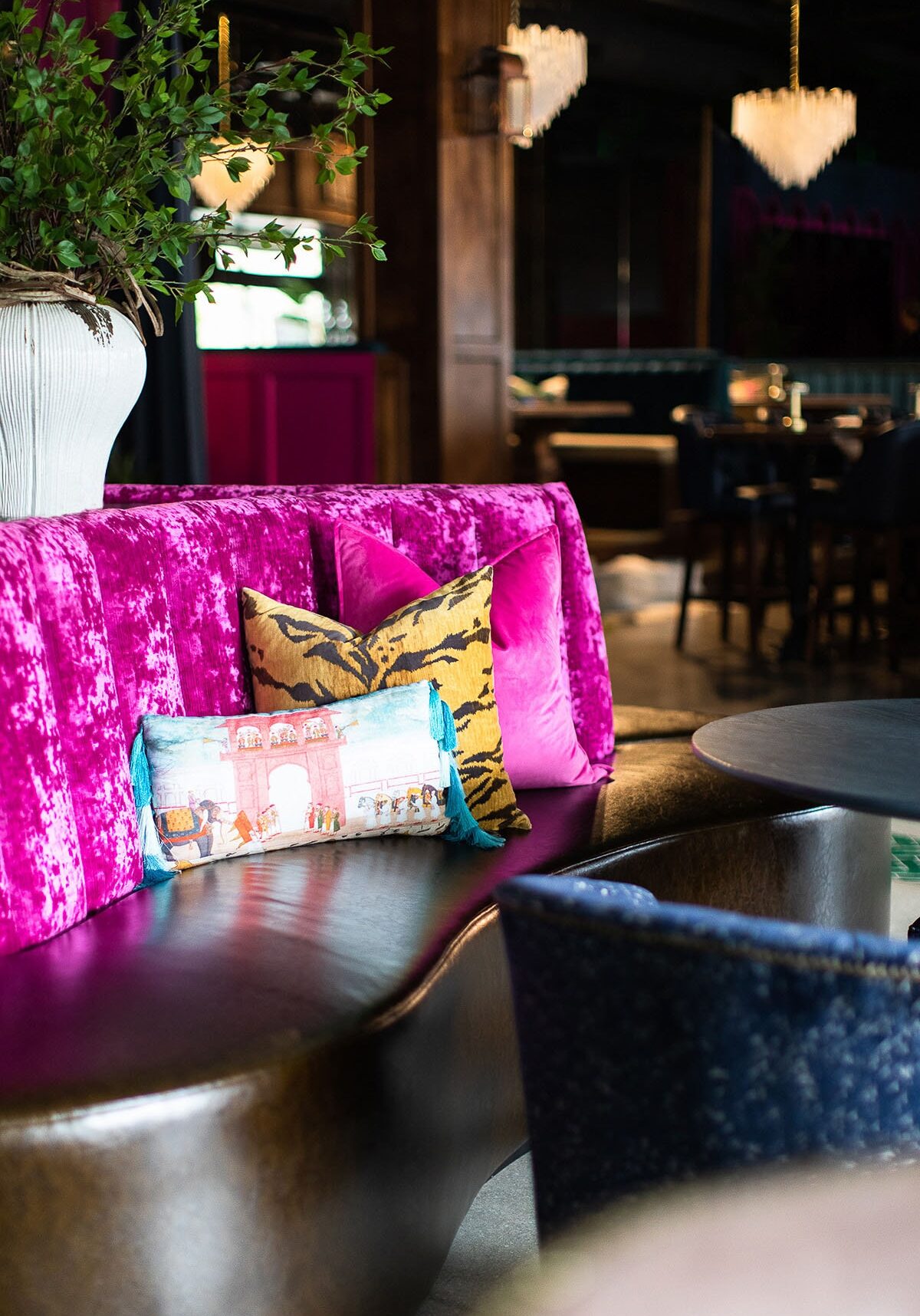 A plush, magenta velvet booth with decorative pillows, including a tiger print and a tasseled cushion, in a stylish, upscale lounge with chandelier lighting.