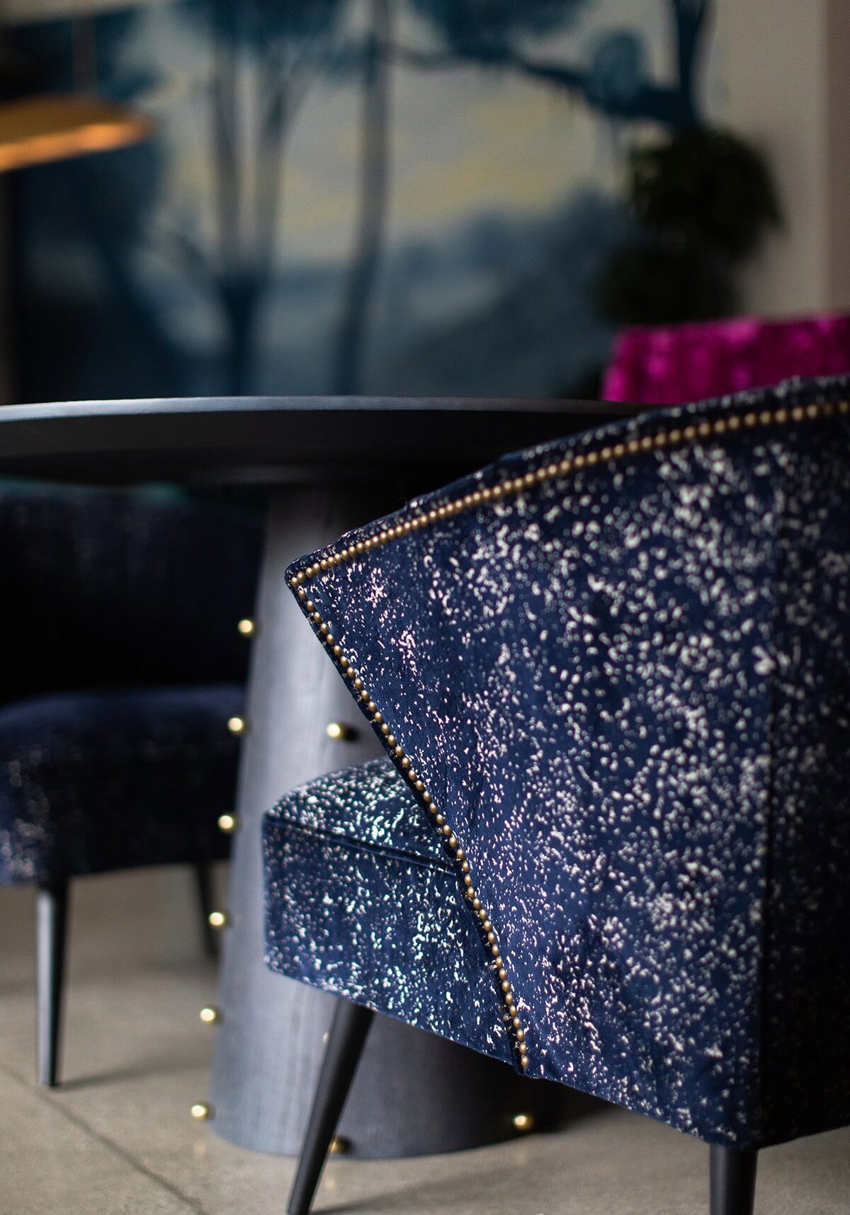Close-up of a blue velvet chair with gold studs next to a round dark table in a modern interior setting.
