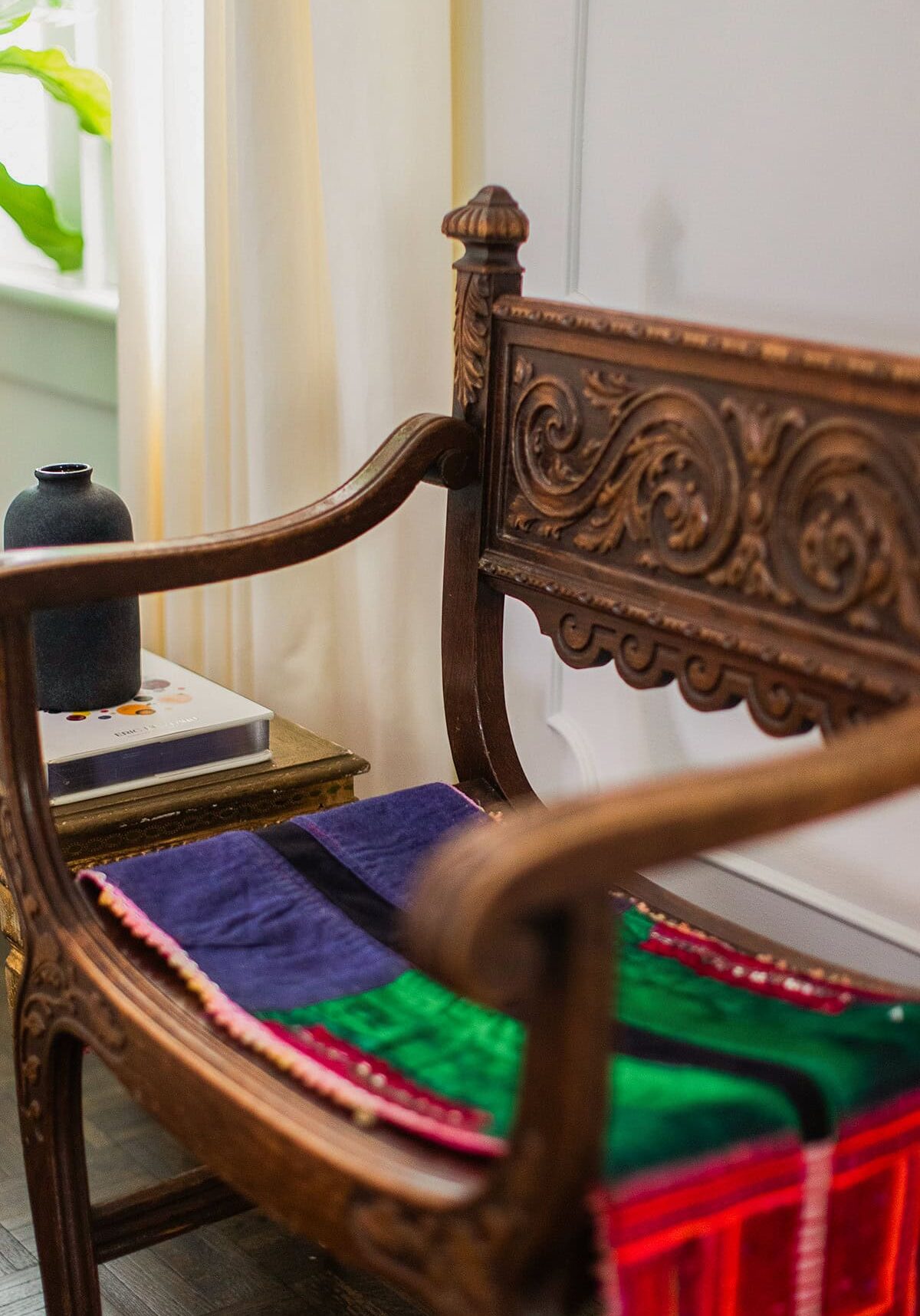 A carved wooden chair with a colorful, striped cushion sits beside a small table holding a black vase and a book, near a window with light-colored curtains.