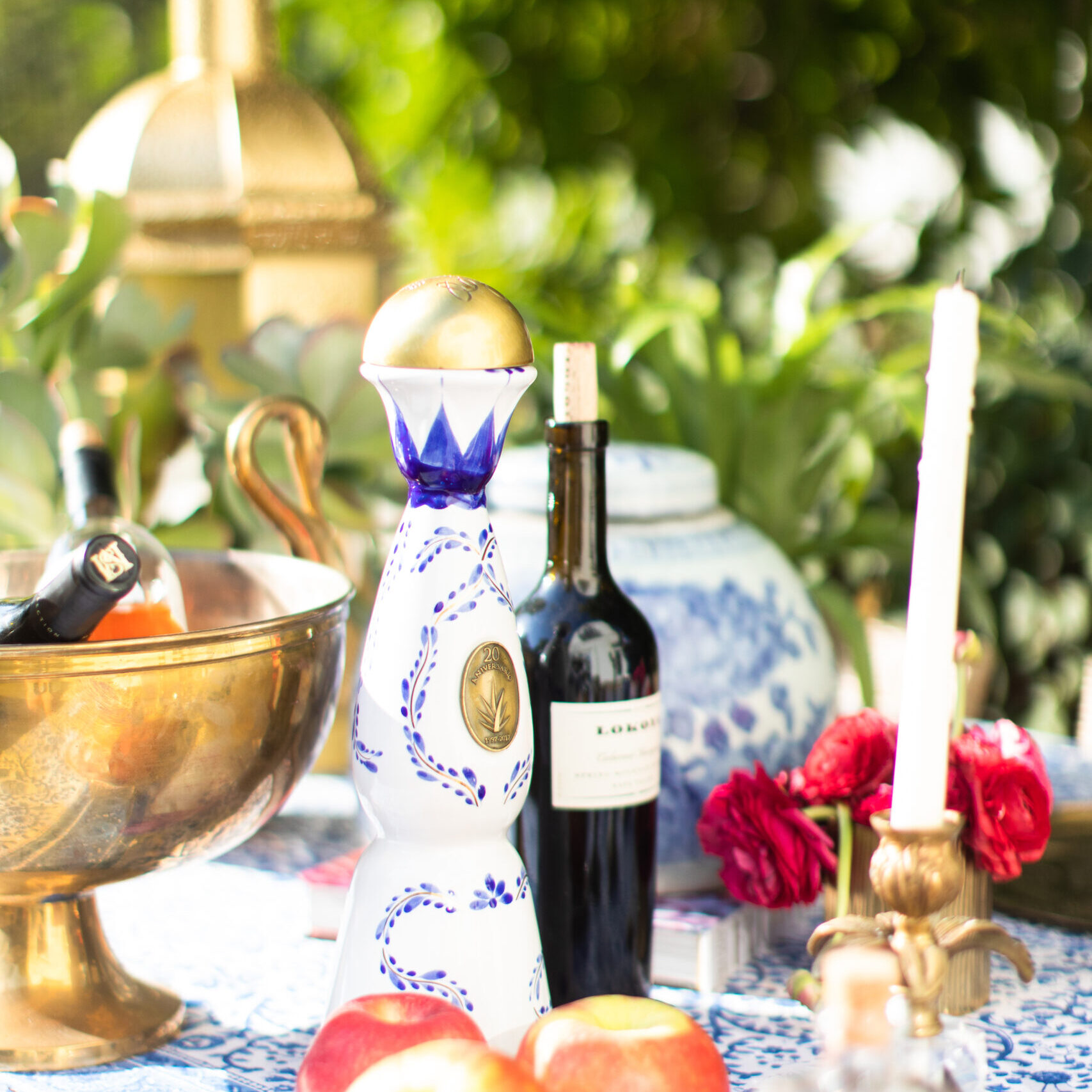 A bottle of tequila, a bottle of red wine, apples, a candle, and floral decor by Crimson Design Group are artfully arranged on a patterned table outdoors.