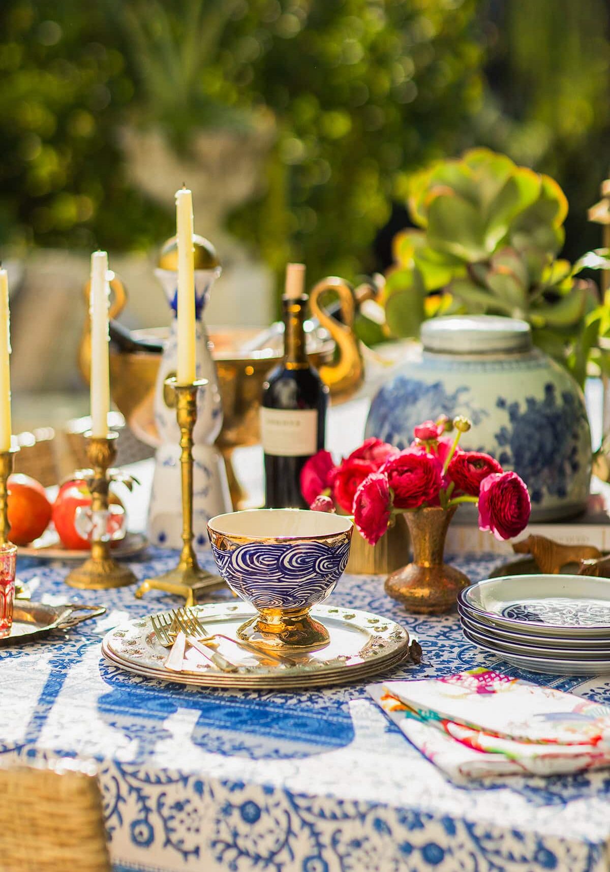 A colorful outdoor table setting with patterned plates, gold cutlery, candles, a bowl with a blue design, red flowers in a vase, and a wine bottle on a blue and white tablecloth.