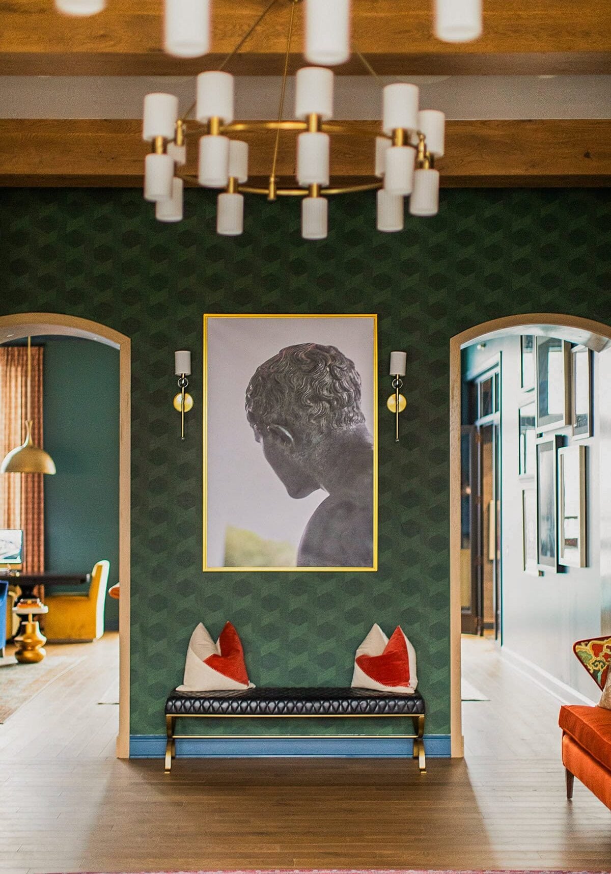 A hallway with green patterned walls features a large framed sculpture photo, a bench with red and white cushions, and a modern chandelier overhead.
