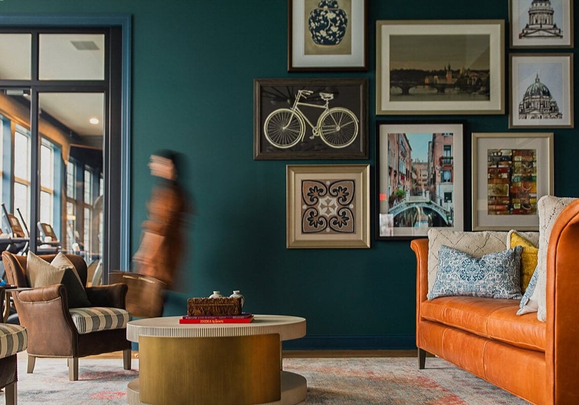 A blurred person walks in a modern living room with a green wall, leather chairs, a round coffee table, and framed art including a bicycle and cityscape.