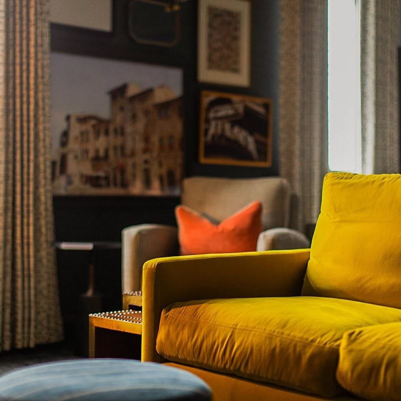 A room with a yellow sofa, a plaid pillow, an armchair with an orange pillow, patterned curtains, framed artwork, and soft natural light from the windows.