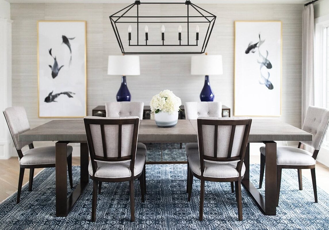 A modern dining room with a wooden table, six cushioned chairs, two blue lamps, floral centerpiece, abstract art, and a geometric chandelier over a blue patterned rug.