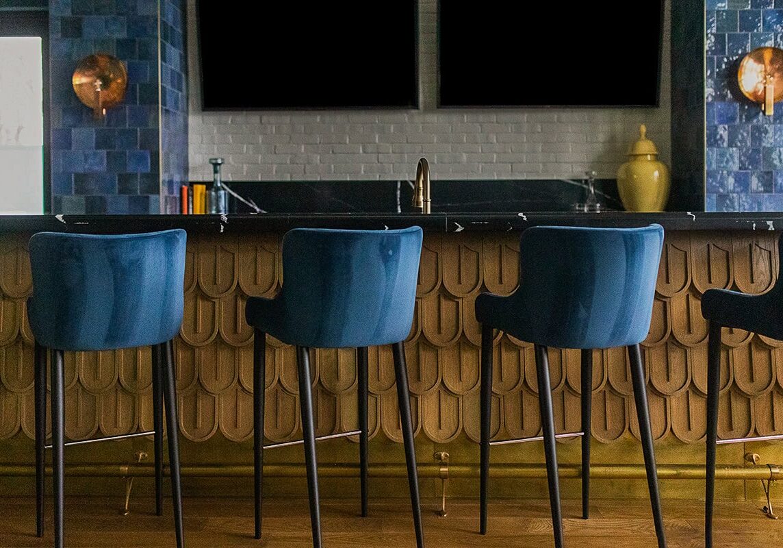 Four blue barstools are lined up at a bar with a patterned front panel; the bar area features blue tiles, wall sconces, and a white brick backsplash.