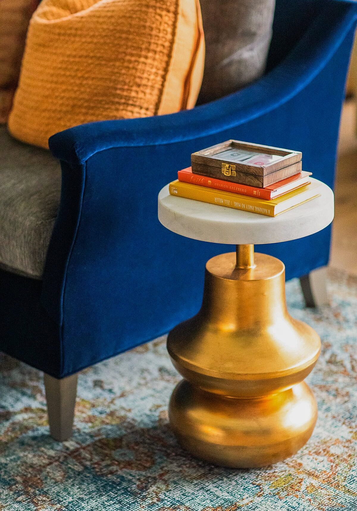 A round gold side table with a white top holds stacked books and a wooden box, positioned next to a blue upholstered chair with an orange pillow.