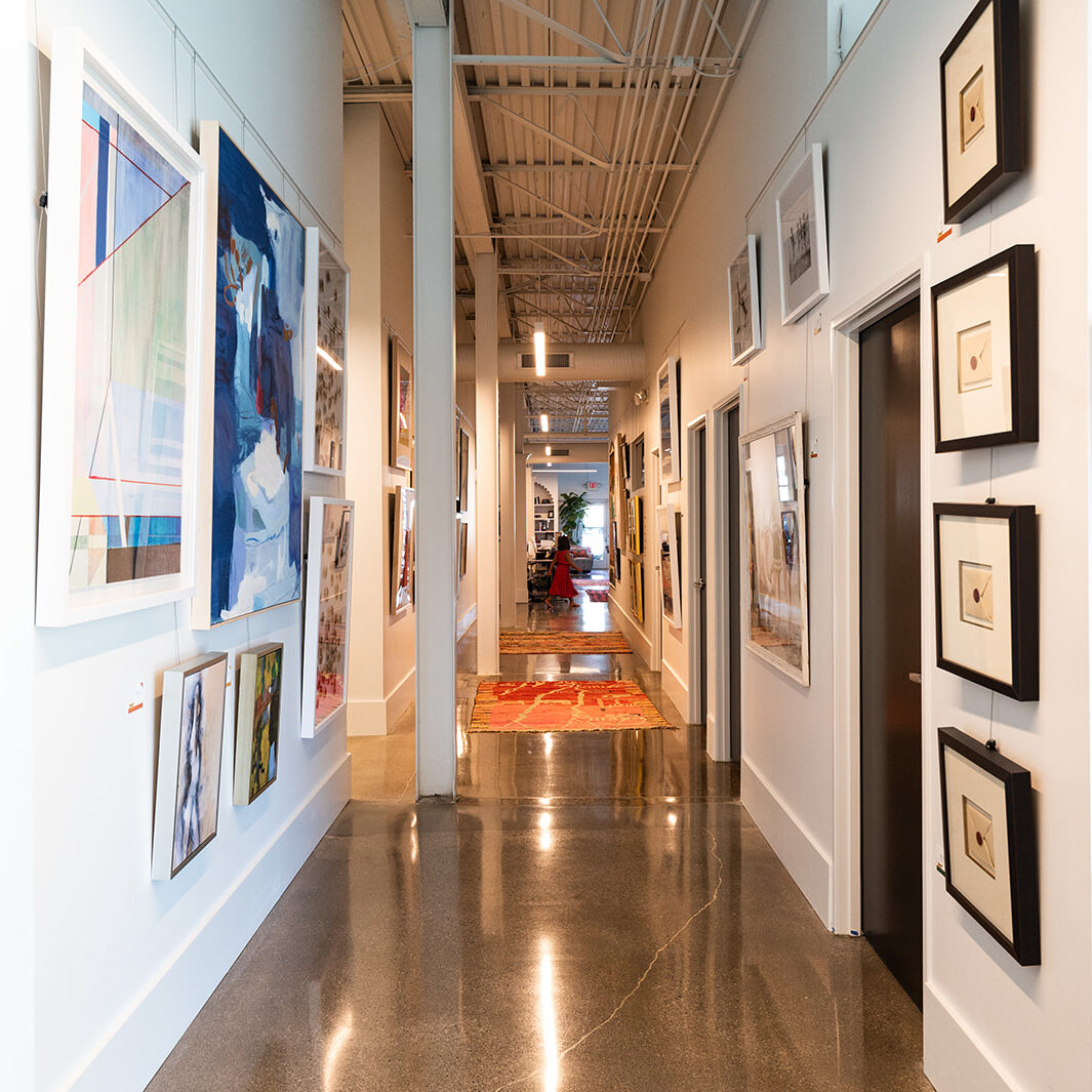 A brightly lit hallway with polished floors, designed by Crimson Design Group, is lined with framed artwork on white walls and leads to a distant room with red chairs and a plant.