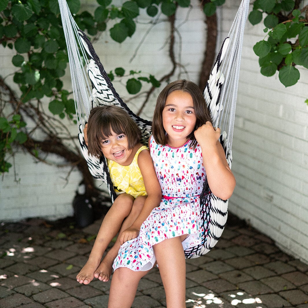 Two young children sit together on a hammock chair outdoors, both smiling at the camera, surrounded by greenery and a white brick wall—a charming scene inspired by Crimson Design Group style.