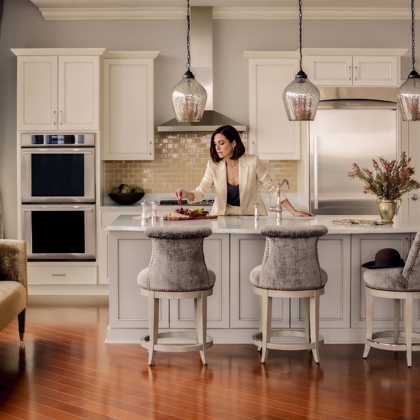A woman in a cream blazer stands at a kitchen island, arranging grapes on a serving board.