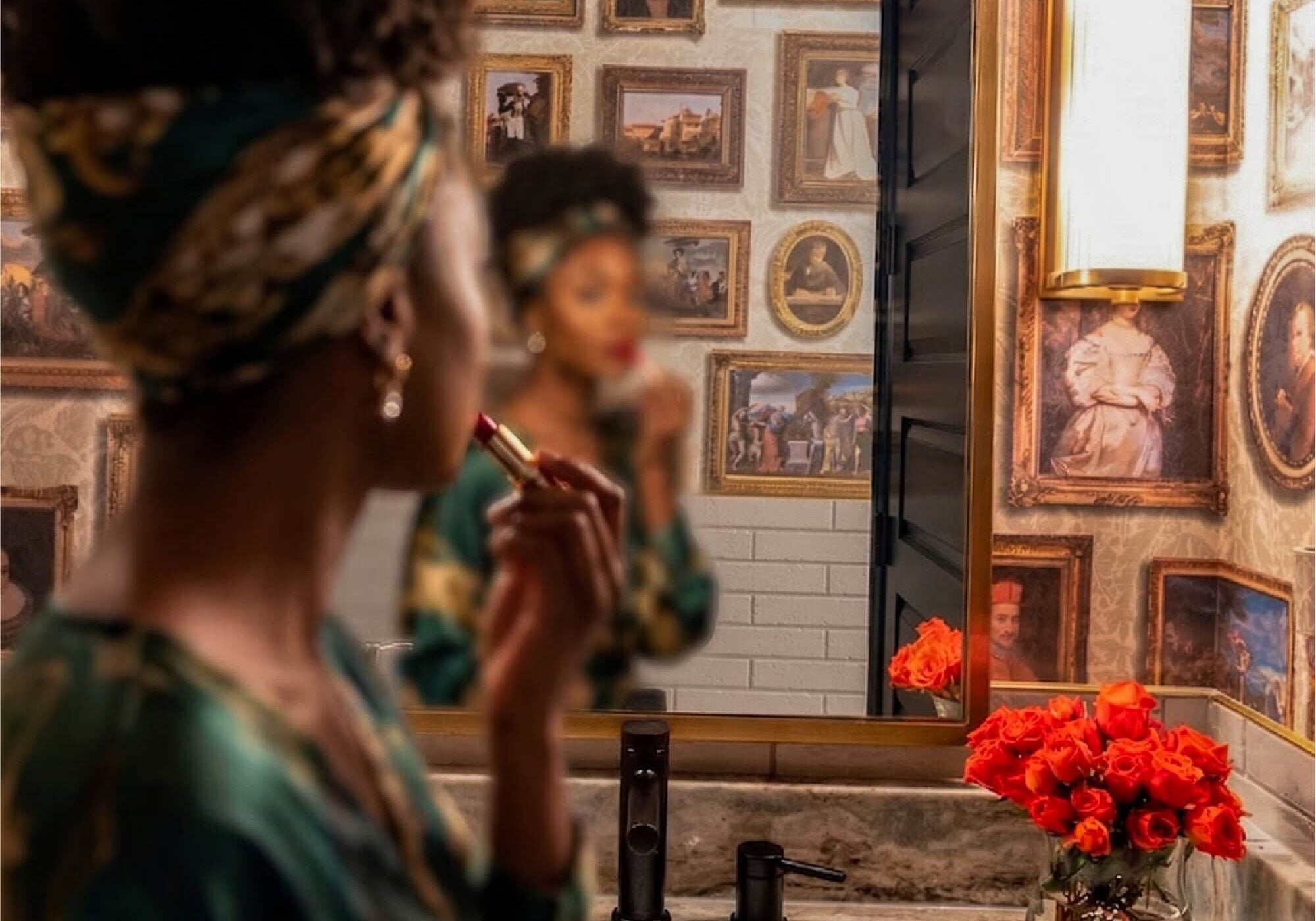A person applies lipstick in front of a bathroom mirror, with framed artwork on the wall and a vase of red roses on the marble countertop.