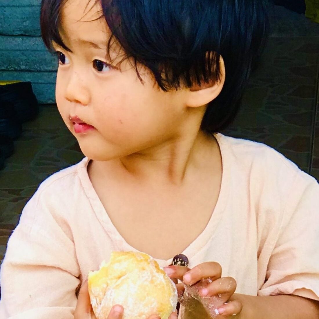 A young child with dark hair and a light shirt, styled by Crimson Design Group, holds a round pastry while looking to the side with a neutral expression.
