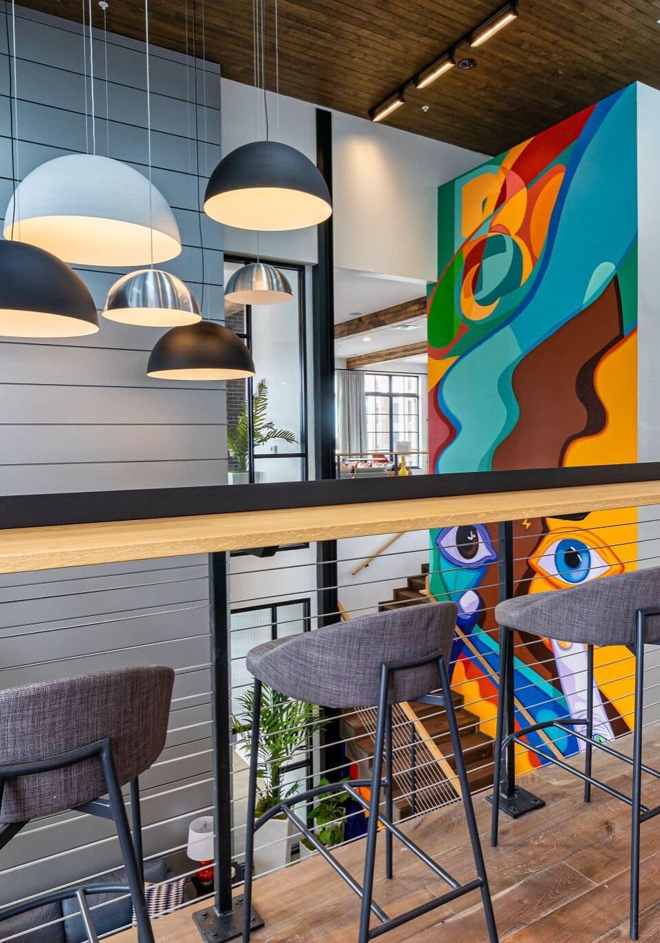 Modern office lounge with high chairs, wooden floors, pendant lights, and a colorful abstract mural on the far wall. Large windows provide ample natural light.
