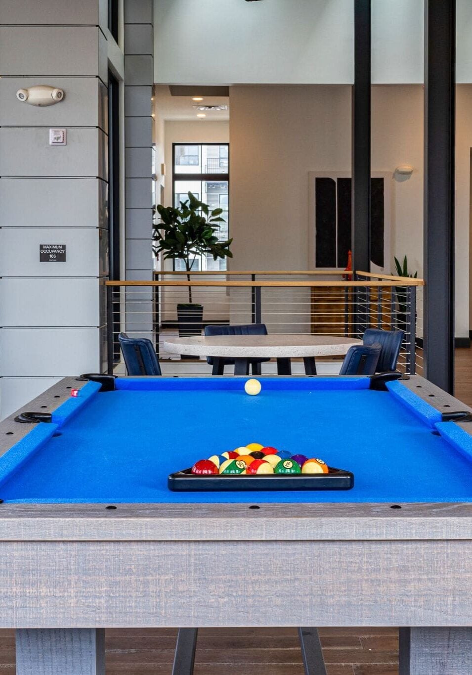 A blue pool table with a rack of balls and a cue ball stands in the center of a modern, open lounge area with wooden floors and contemporary furnishings.