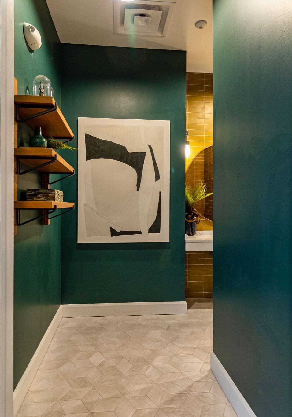 Entrance to a men's restroom with green walls, a black-and-white abstract painting, shelves with decor, and a glimpse of a sink and mirror inside.