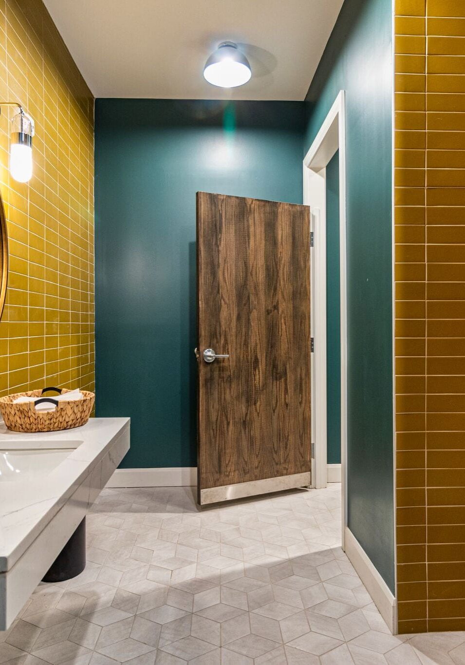 A modern bathroom with yellow tiled walls, dark green painted sections, a wood door, a floating sink with a mirror, and geometric light gray floor tiles.