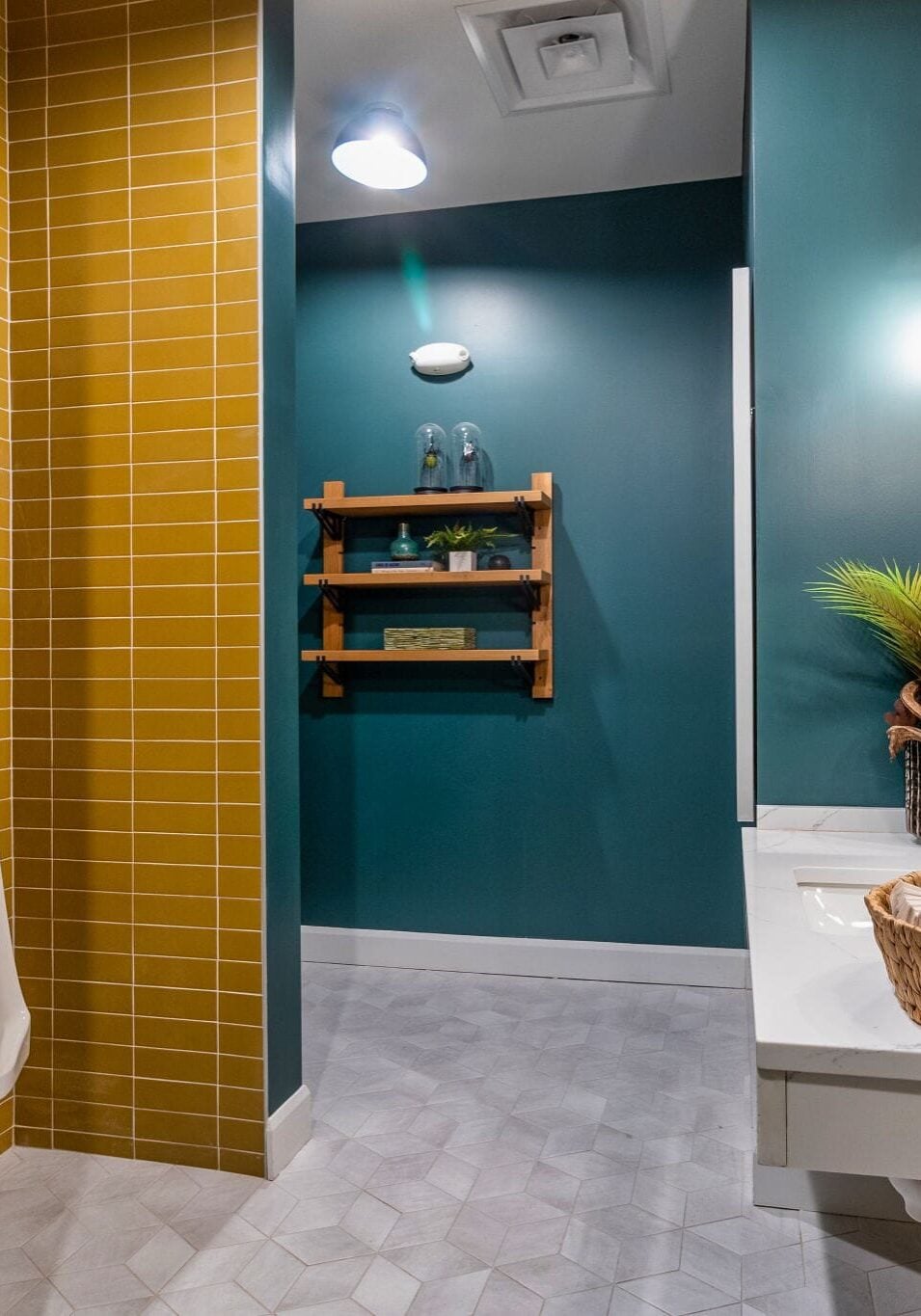 Modern bathroom with teal and mustard yellow walls, a round mirror, double sinks, decorative plants, and a basket on the counter. A wall-mounted shelf and urinal are visible in the background.