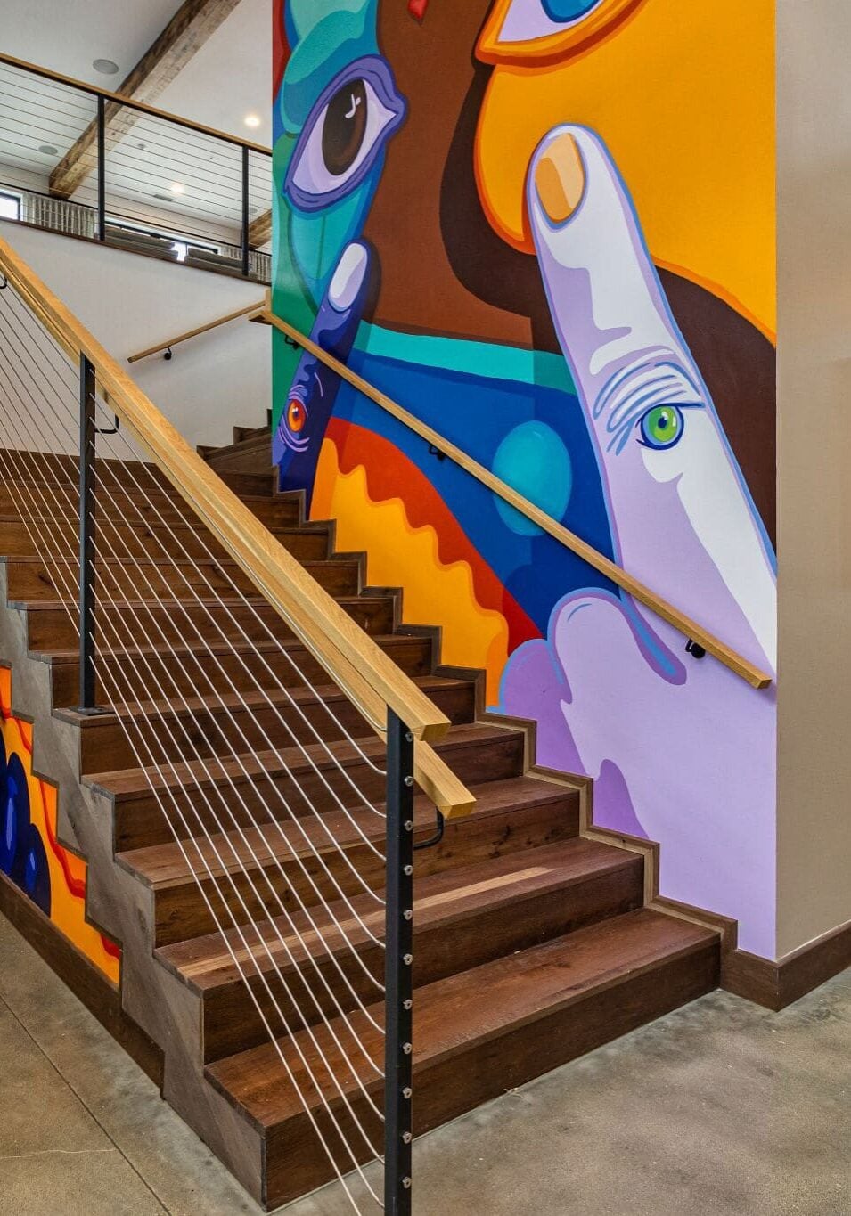 A modern indoor staircase with a vibrant, abstract mural featuring faces and hands on the adjacent wall; potted plants are placed nearby.
