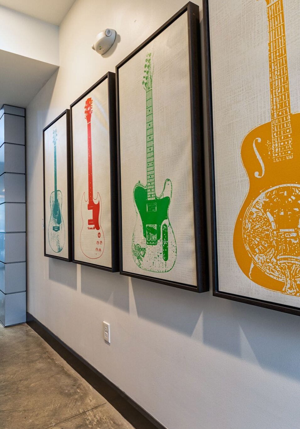 A hallway with concrete floors and framed colorful guitar artwork on the wall; modern office spaces are visible in the background.
