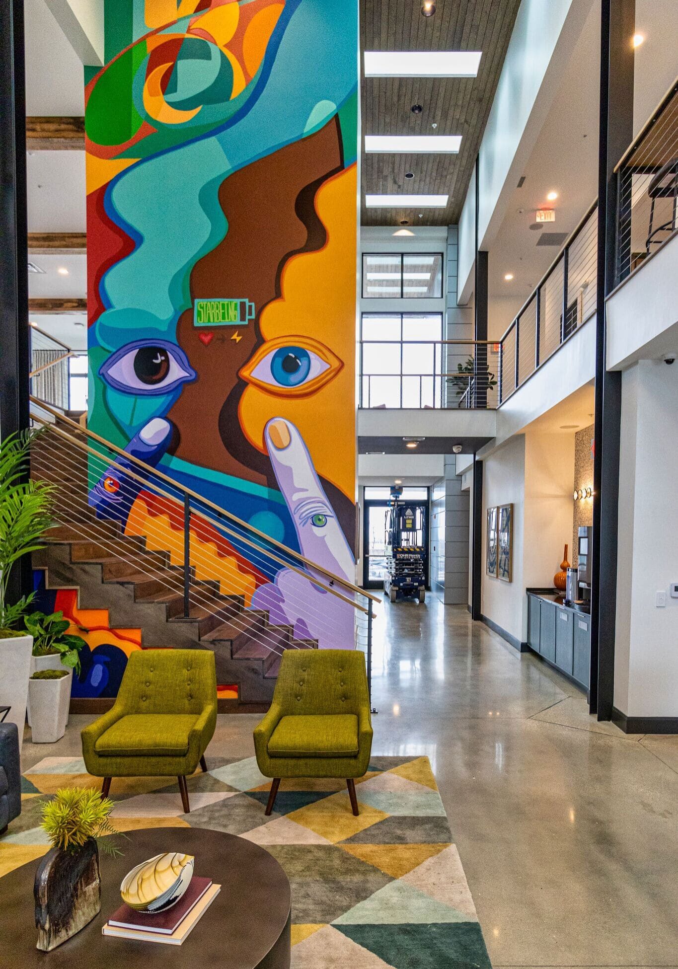 A modern lobby with green chairs, a round table, and a colorful abstract mural featuring faces on a tall wall by a staircase.