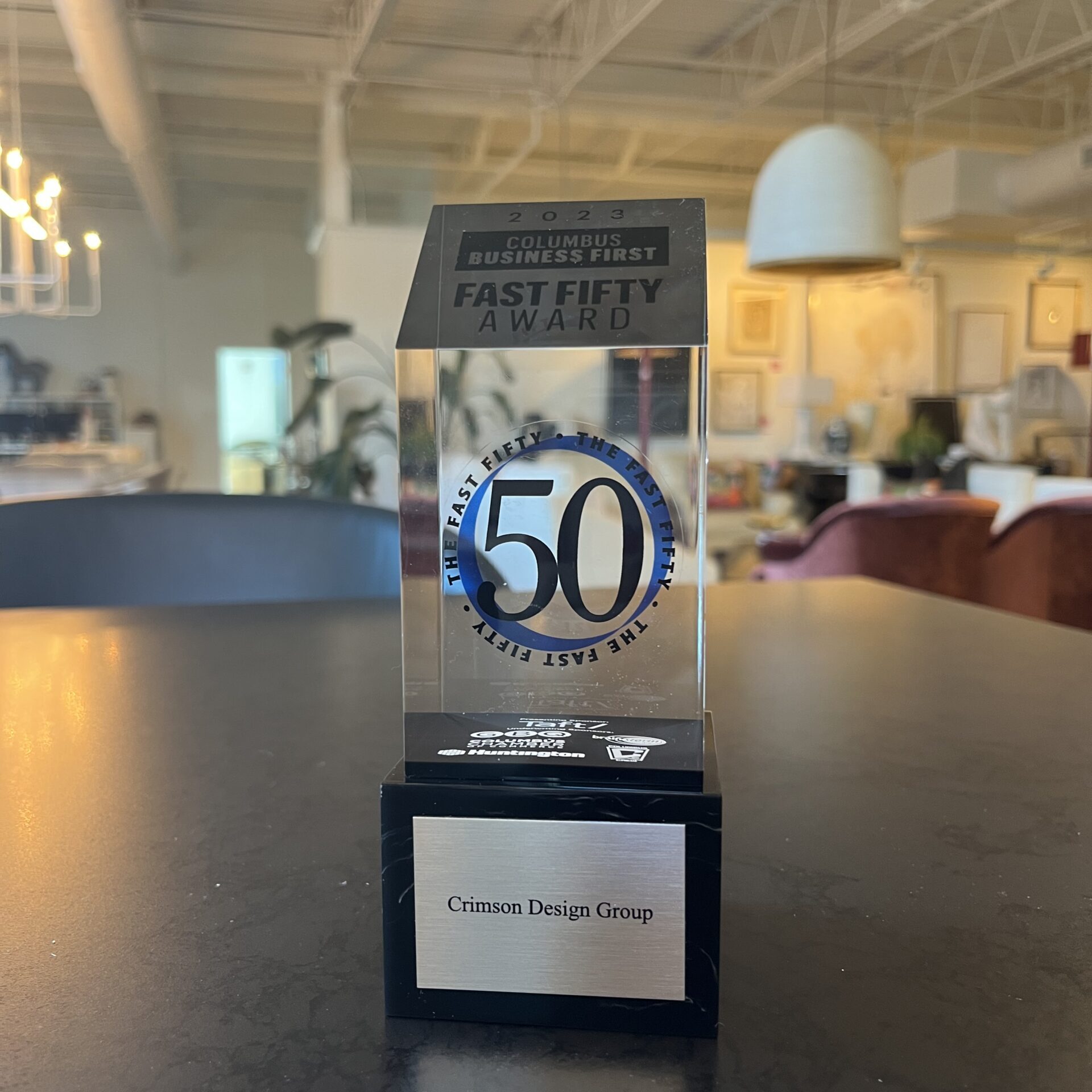 A glass trophy labeled "Fast Fifty Award" sits on a table in an office, proudly displaying "Crimson Design Group" at its base, celebrating the company’s outstanding achievement.