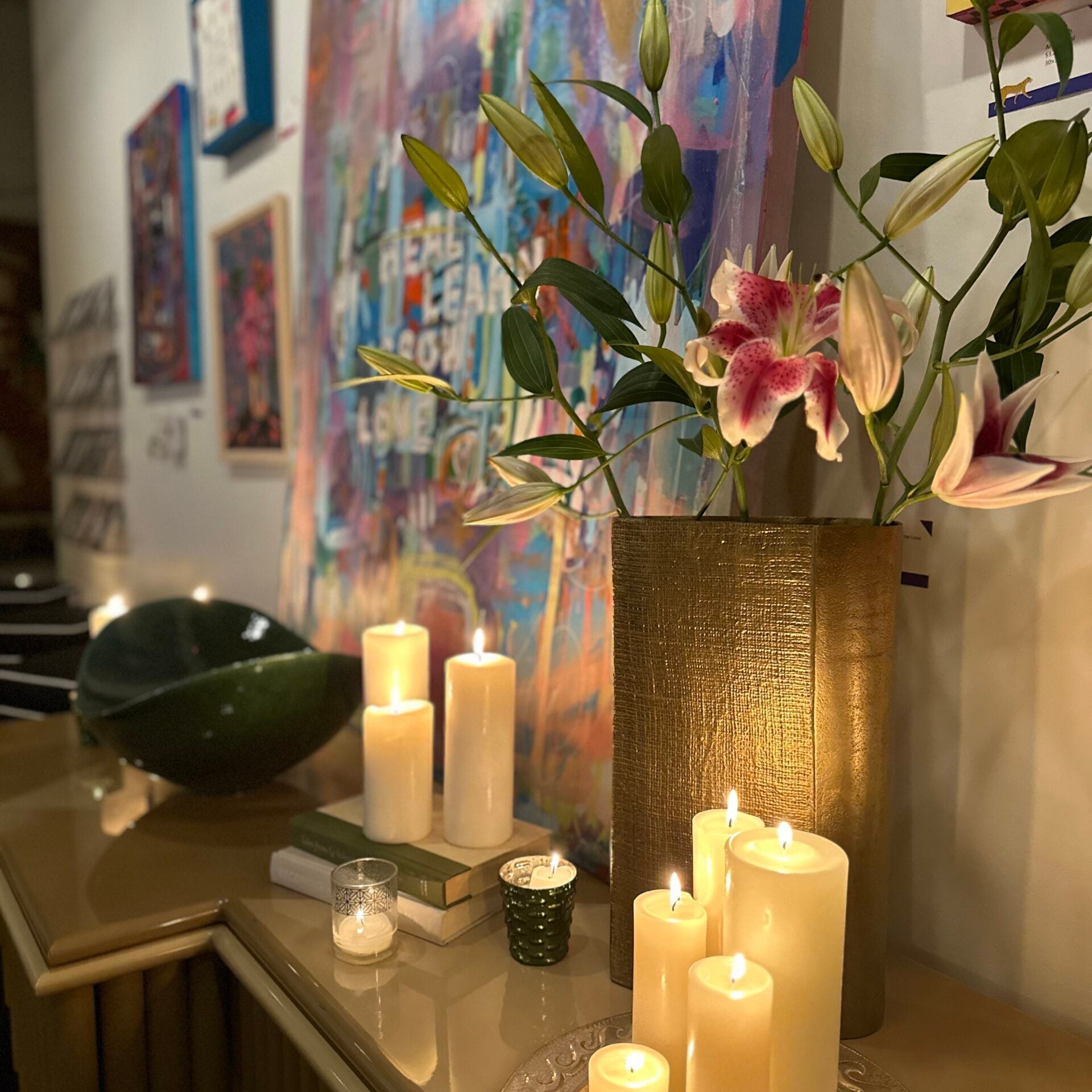 A gold vase with lilies and several lit candles are arranged on a table in front of colorful abstract paintings, showcasing Crimson Design Group's signature vibrant style.
