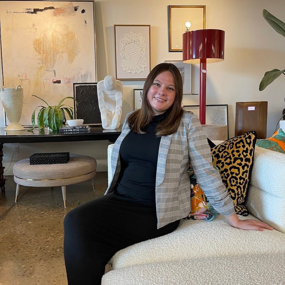 A woman in a plaid blazer sits on a white couch in a modern, art-filled living room with contemporary decor and wall art by Crimson Design Group.