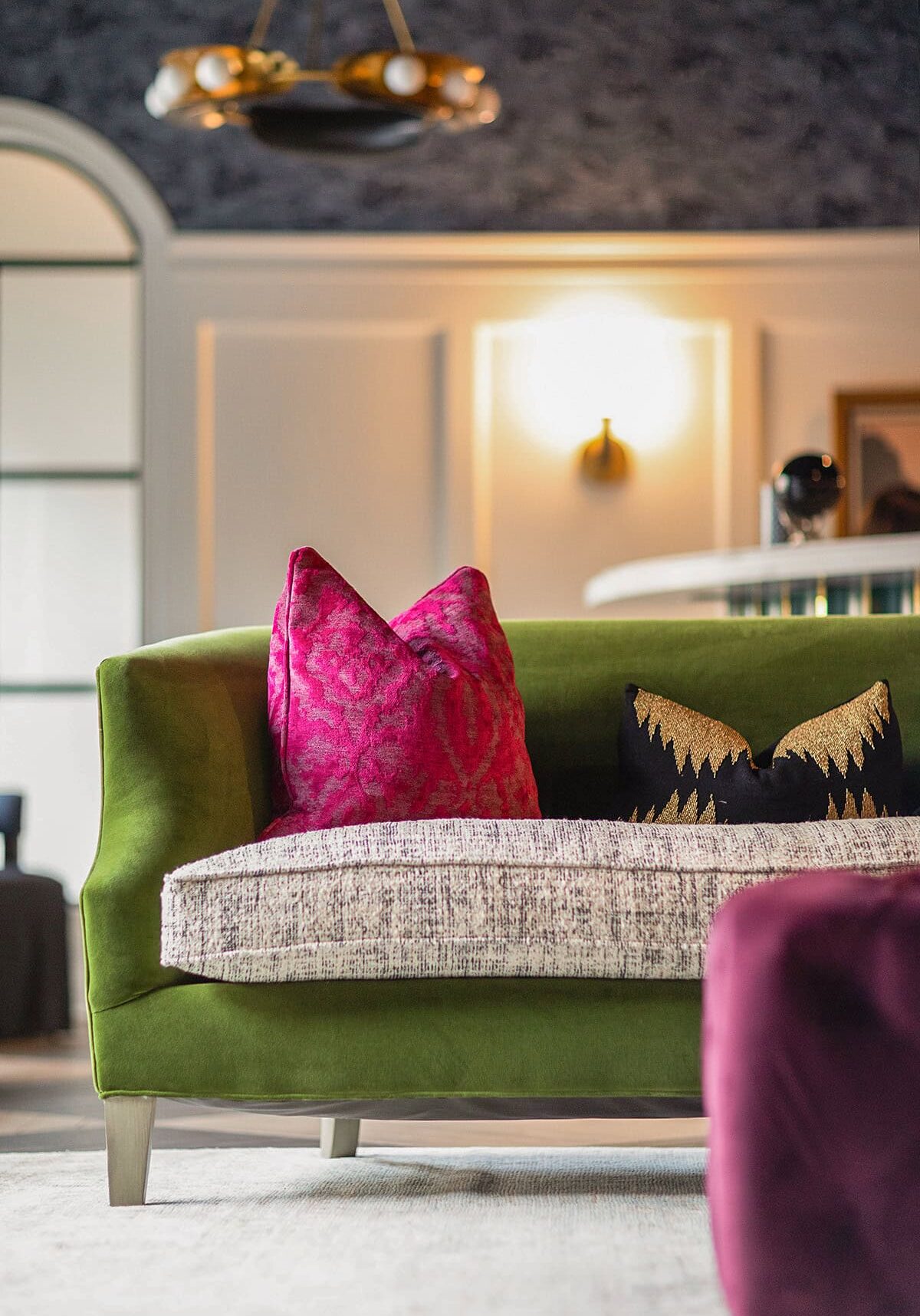 Green sofa with textured seat cushion, two accent pillows—one bright pink and one black with gold pattern—sits in a modern, elegantly decorated room.