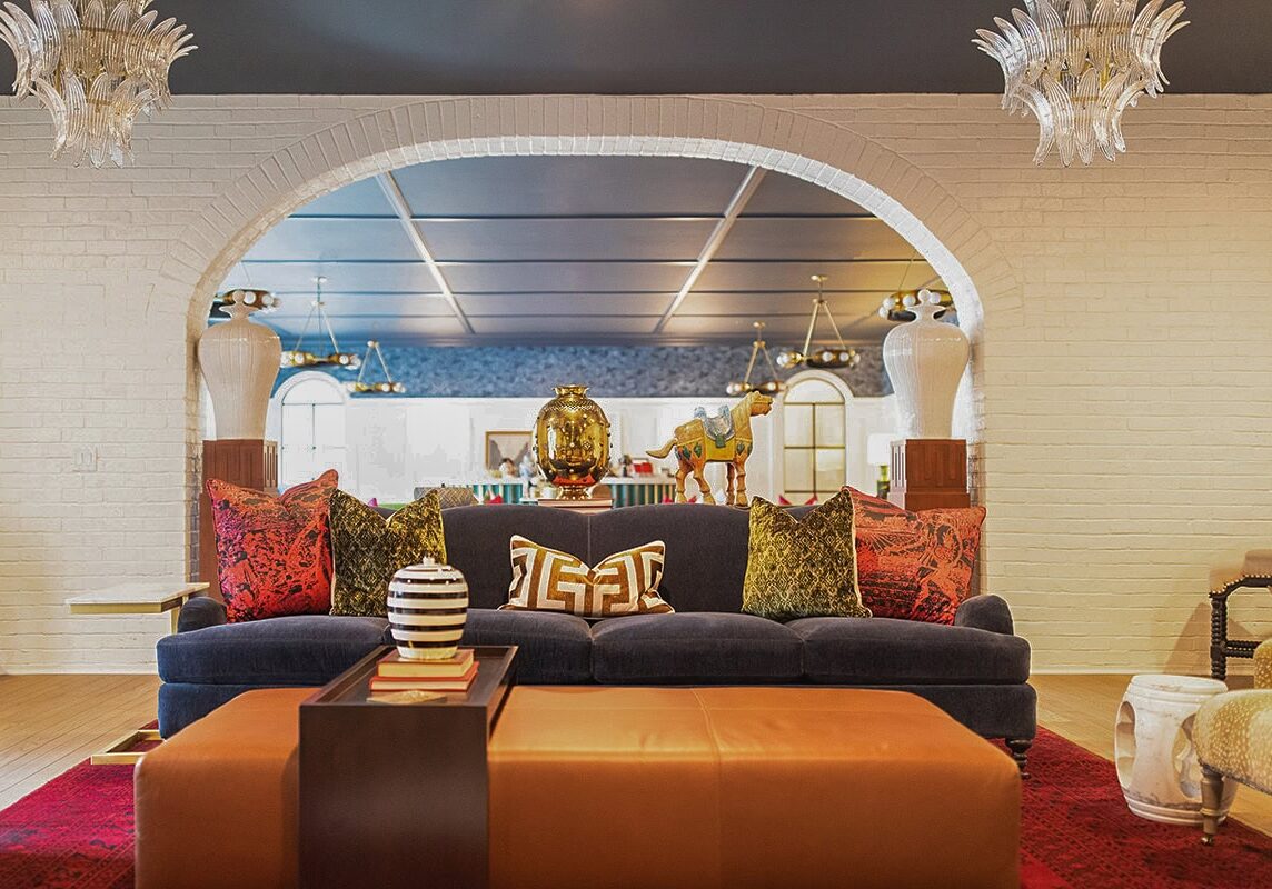 A living room with a blue velvet sofa, colorful patterned pillows, a camel figurine, and a gold vase, set under a white brick archway with modern chandeliers overhead.