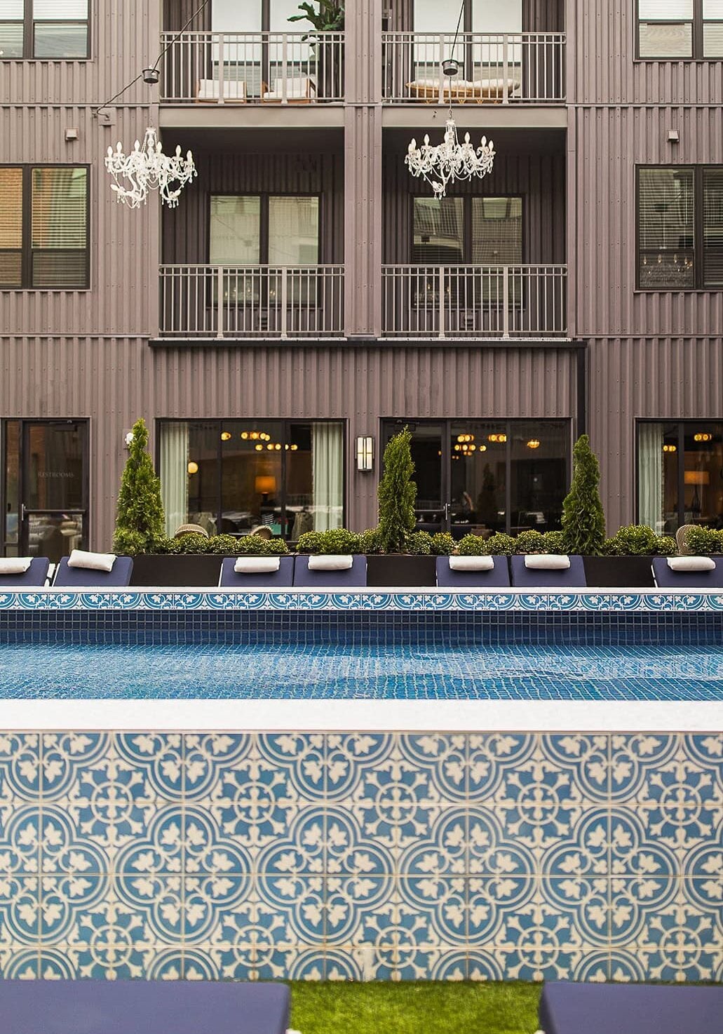 A patterned tile swimming pool with blue lounge chairs, in front of a modern building with balconies and hanging chandeliers.