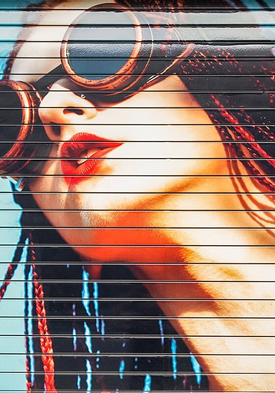 Colorful mural of a woman with red braids, wearing large round goggles and red lipstick, painted on a corrugated metal surface against a bright blue background.